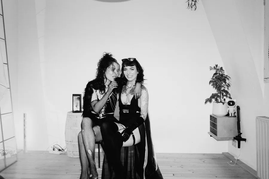 Black and white: two women in black outfits sitting close together in a bright room with a plant, flash by Kilian Amrehn