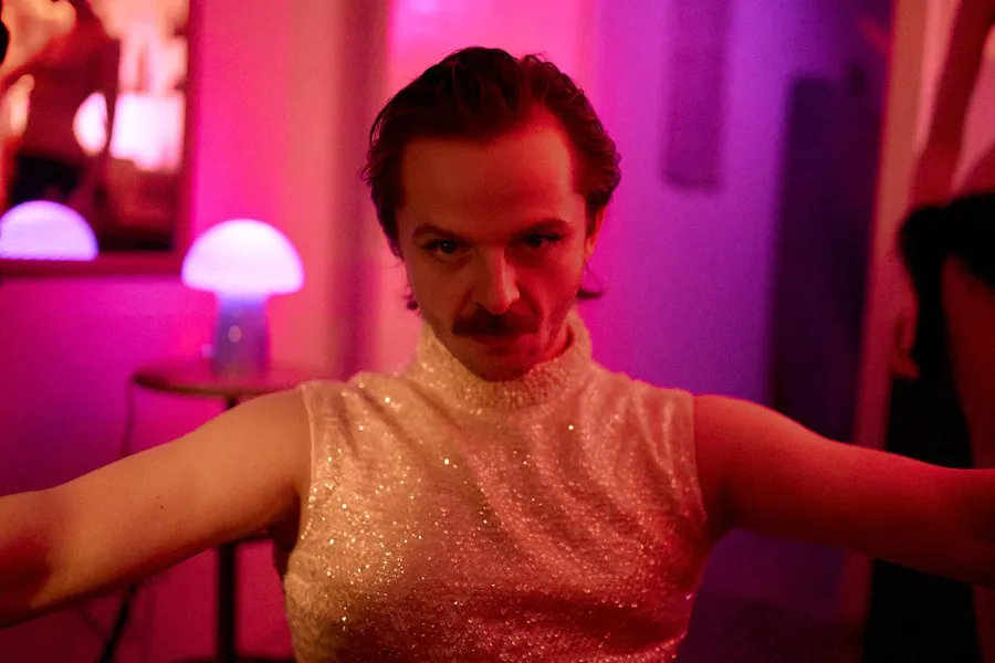 Man with a moustache in a glitter top spreading his arms, pink and purple party light, mushroom lamp, flash by Kilian Amrehn