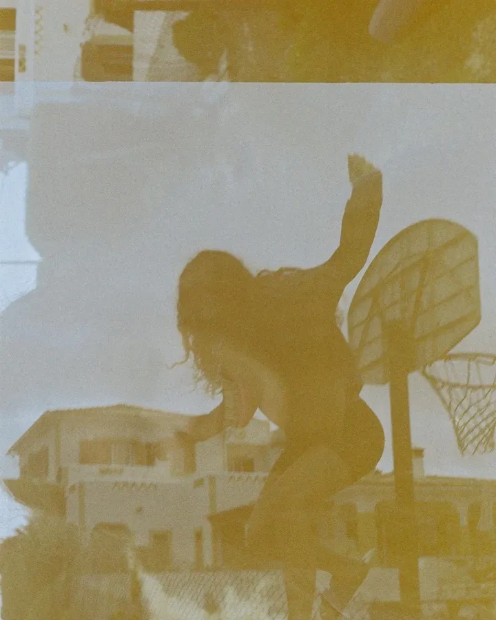 Analogue with a yellow cast: silhouette of a person jumping at a basketball hoop in front of houses, flash by Kilian Amrehn