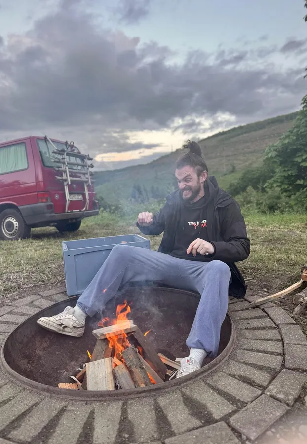 Man sitting laughing at a fire pit, red van and hills at dusk, flash by Kilian Amrehn
