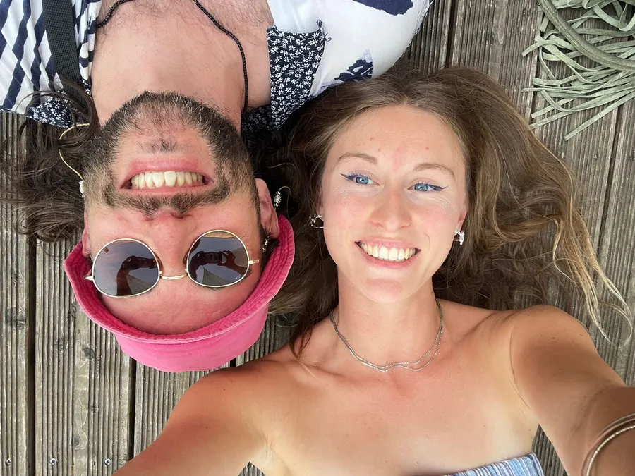 Selfie on a wooden deck: upside-down man with pink hat and round sunglasses, smiling woman, flash by Kilian Amrehn