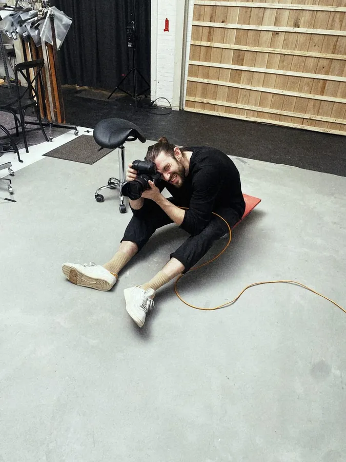 Photographer sitting on a grey studio floor shooting with a camera, cable beside him, flash by Kilian Amrehn