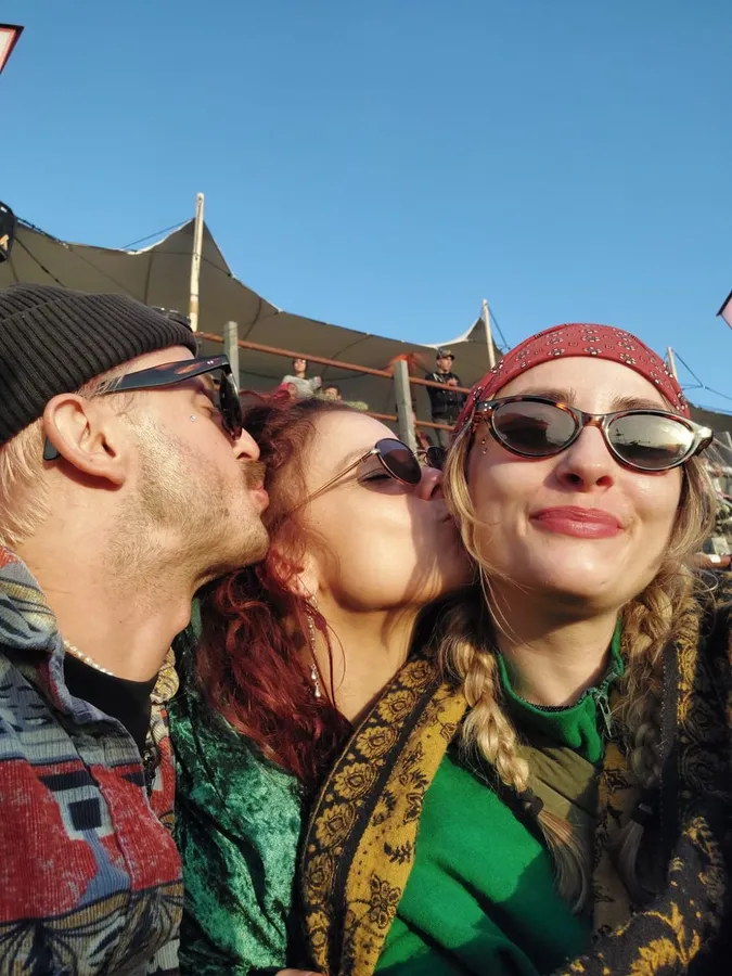 Three friends in sunglasses, a kiss on the cheek, blue sky and tent roof, flash by Kilian Amrehn