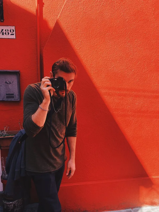 Man taking a photo with a camera in front of a bright red wall with hard shadow, flash by Kilian Amrehn