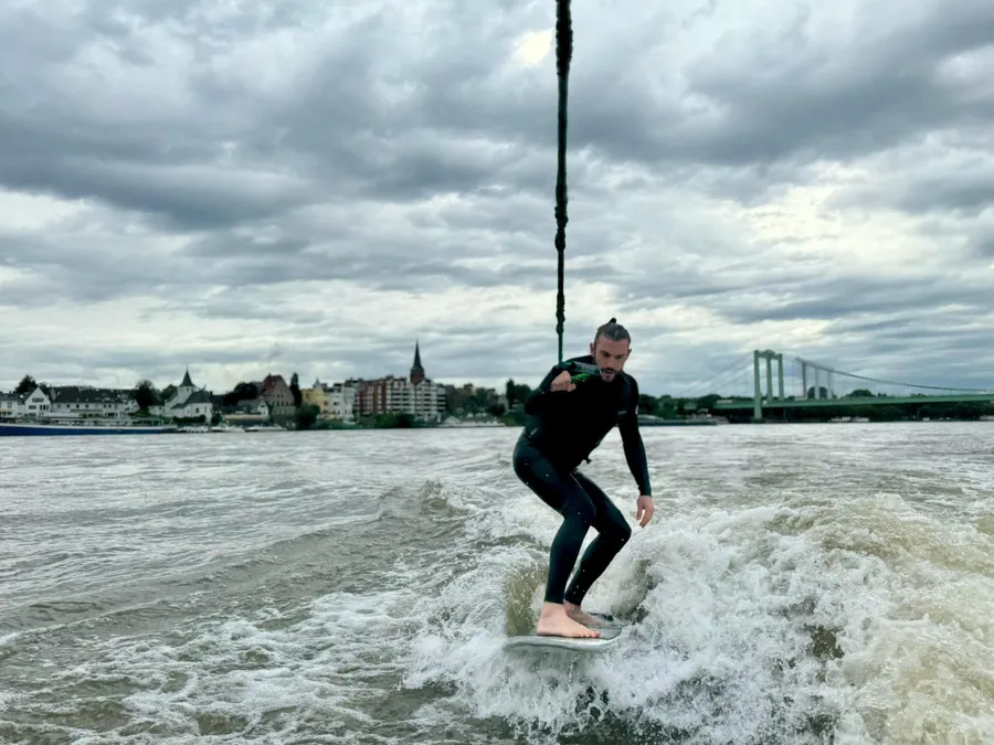 Wakesurfer in a wetsuit on a wide river, church spire and bridge under clouds, flash by Kilian Amrehn