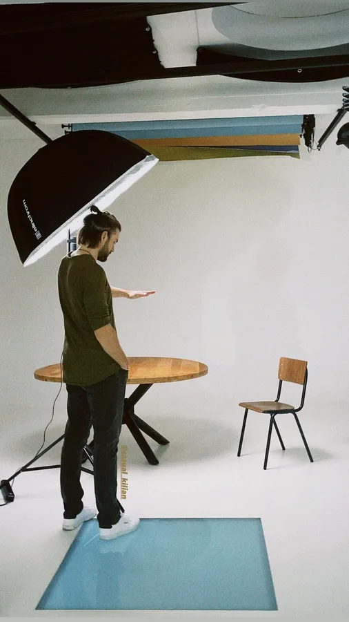 Photographer in a white studio with umbrella light, wooden table and chair as set, flash by Kilian Amrehn