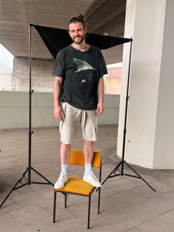 Man standing laughing on a chair in front of a black backdrop under a bridge, flash by Kilian Amrehn