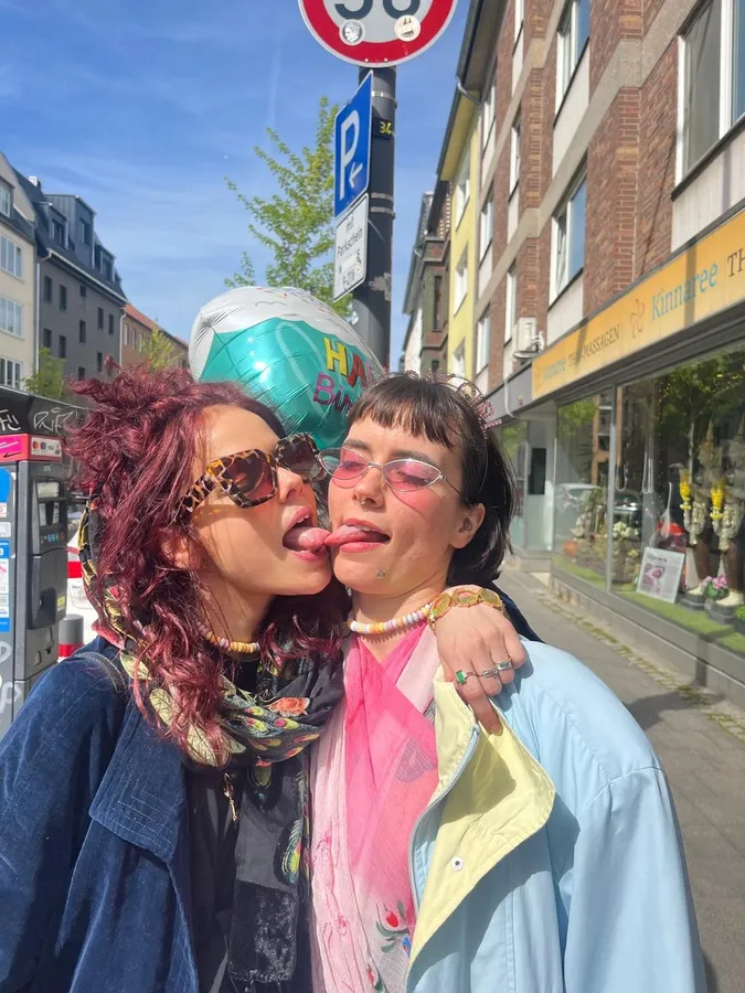 Two women playfully sticking out their tongues at each other on a sunny street, flash by Kilian Amrehn