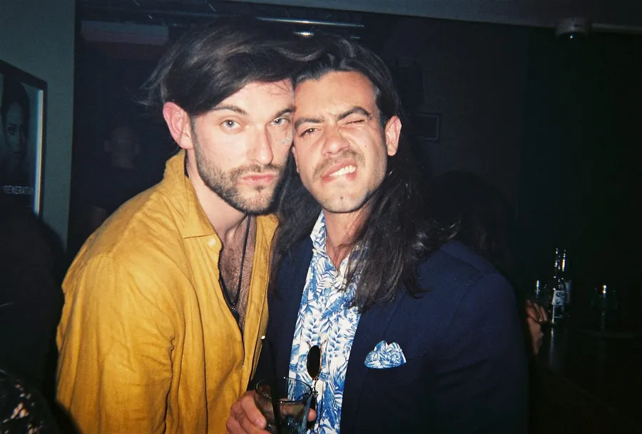 Analogue flash: two men cheek to cheek in a dark bar, mustard shirt and floral shirt, flash by Kilian Amrehn