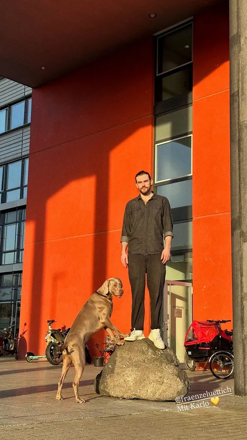 Man standing on a boulder in front of an orange facade, a dog beside him, flash by Kilian Amrehn