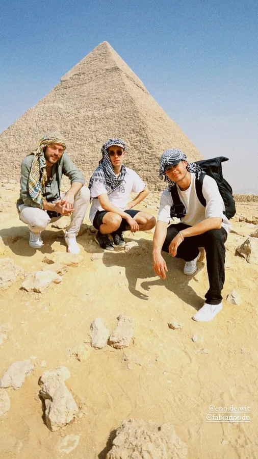 Three men in headscarves crouching in the sand in front of the Pyramid of Giza, flash by Kilian Amrehn