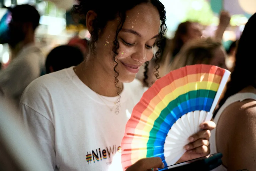 Woman with glitter on her face smiling behind a rainbow fan at a Pride parade, flash by Kilian Amrehn