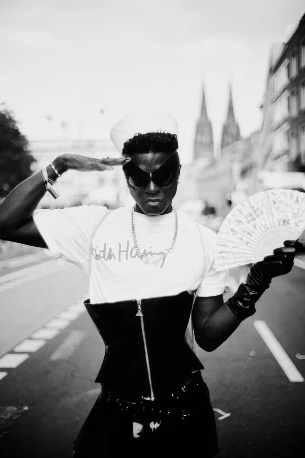 Black and white: person in corset and sailor cap saluting, Cologne Cathedral behind, flash by Kilian Amrehn