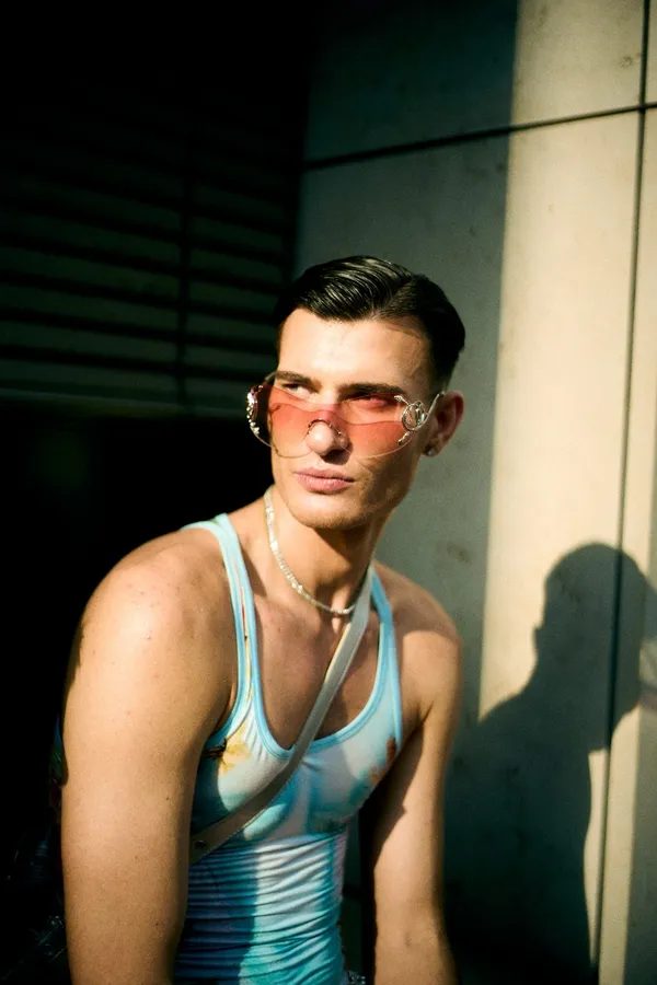 Young man in a tank top and pink sunglasses in hard raking light against a wall, flash by Kilian Amrehn