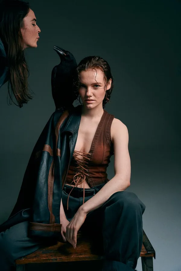 Model in a leather vest seated in a dark studio, a crow perched on a shoulder beside her, flash by Kilian Amrehn
