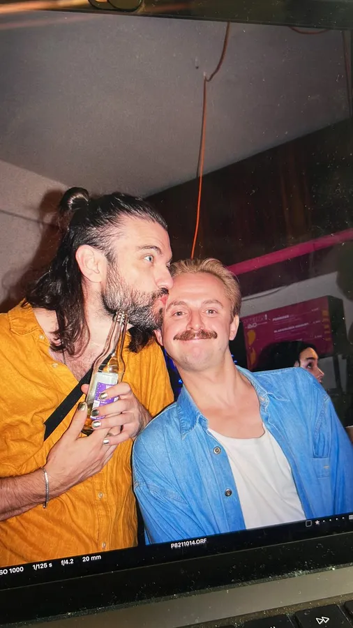 Two men with moustaches, a kiss on the cheek, in front of a laptop on set, flash by Kilian Amrehn