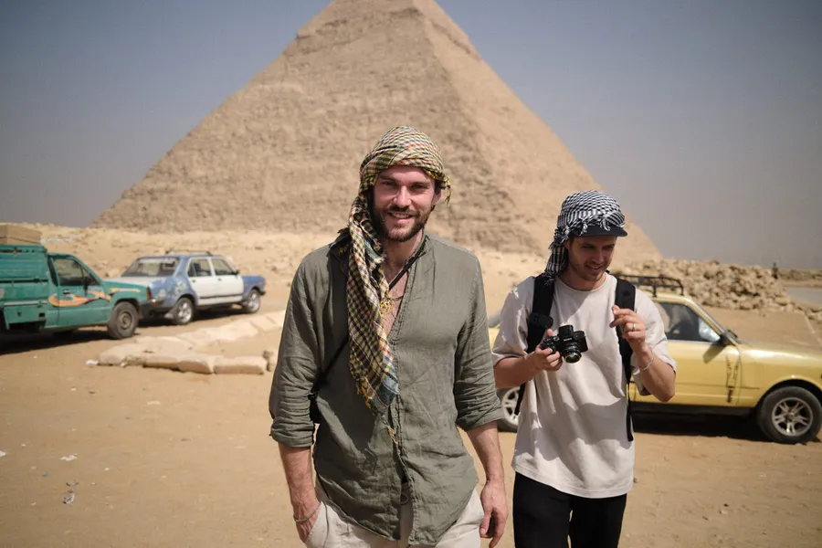 Two men with keffiyehs and a camera in front of the Pyramid of Giza, old cars in the sand, flash by Kilian Amrehn