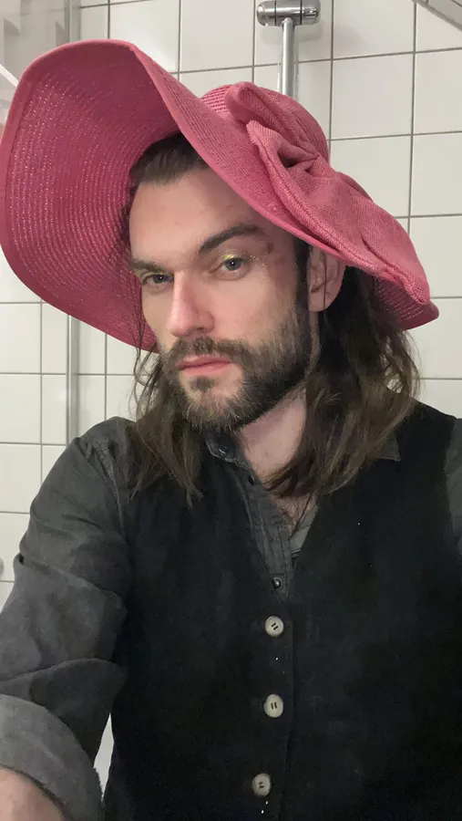 Selfie of a man in a large pink floppy hat against white tiles, flash by Kilian Amrehn