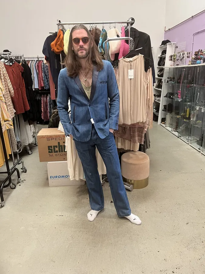 Man in a blue denim suit and sunglasses between clothing rails in a warehouse, flash by Kilian Amrehn
