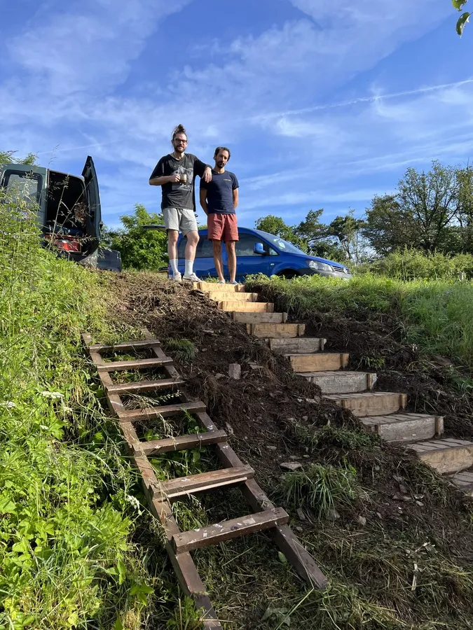 Two men at the top of homemade wooden steps on a slope, blue sky, flash by Kilian Amrehn