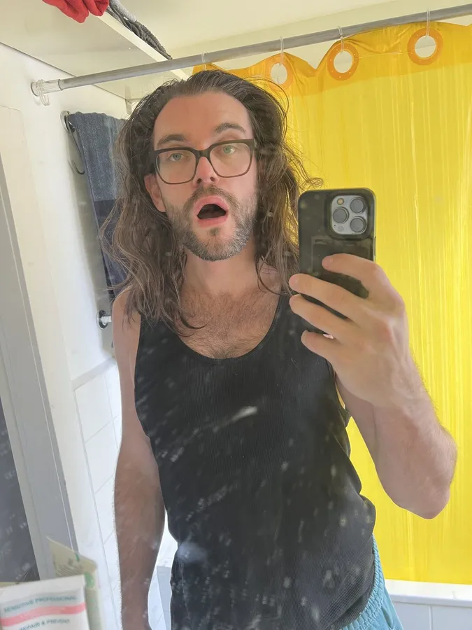 Mirror selfie of a man with glasses and open mouth, yellow shower curtain, flash by Kilian Amrehn