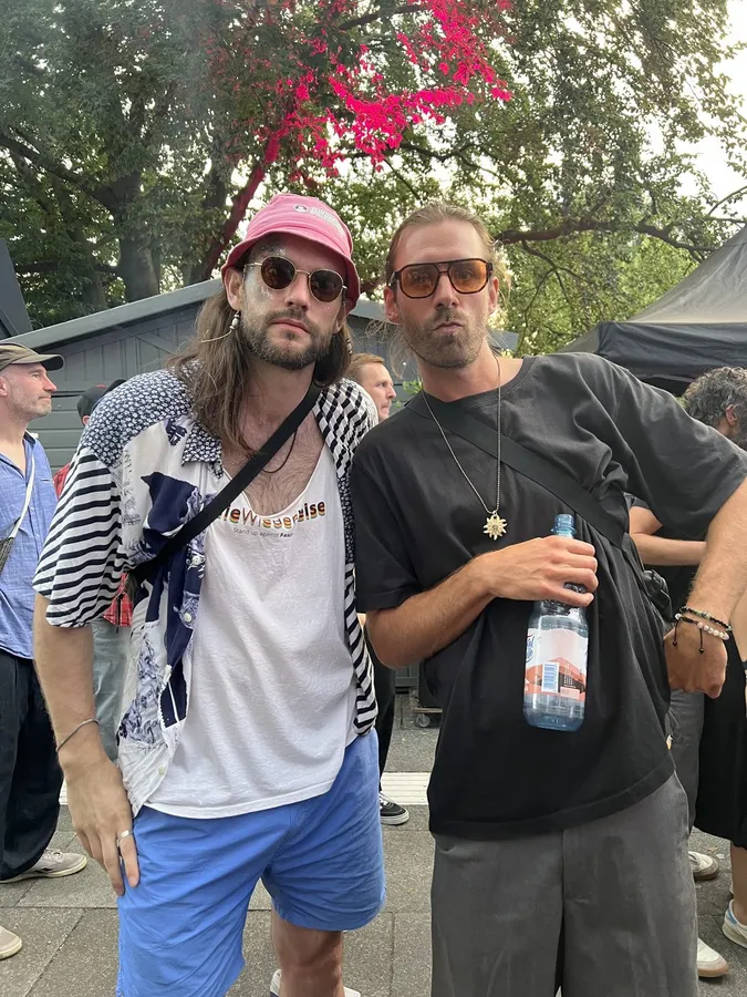 Two men in sunglasses at a street festival, pink hat and water bottle, flash by Kilian Amrehn