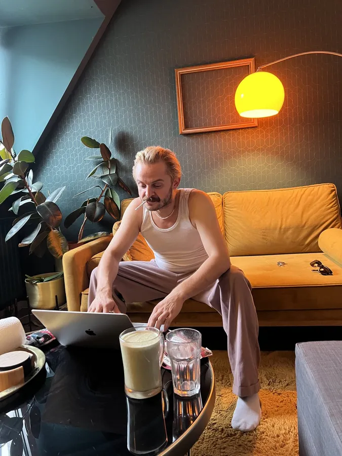 Man in a vest sitting at a laptop in front of a yellow sofa, arc lamp and green wall, flash by Kilian Amrehn