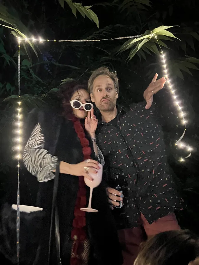 Two people posing at night under fairy lights and palm leaves, white sunglasses, flash by Kilian Amrehn
