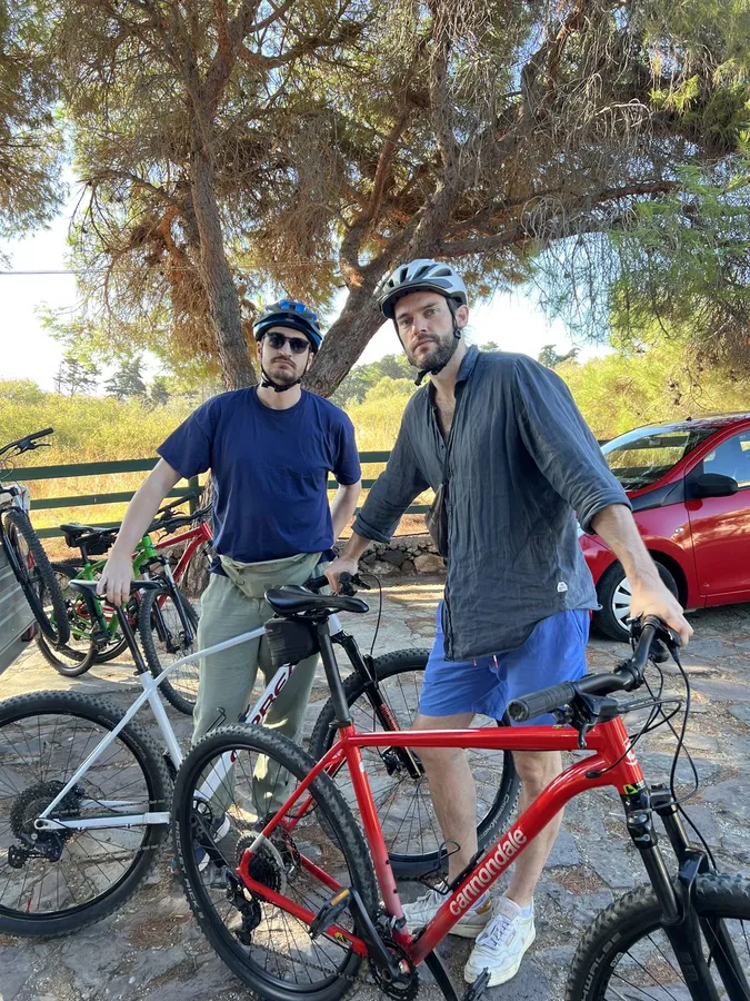 Two men in helmets with mountain bikes under pines, red car behind, flash by Kilian Amrehn