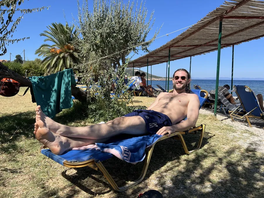 Man lying on a sun lounger under trees by the sea, palm tree and beach shades, flash by Kilian Amrehn