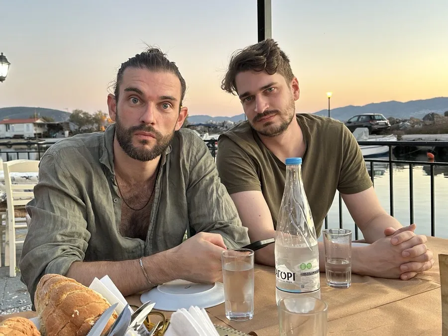 Two men at a harbour taverna table, bread and water, evening sky, flash by Kilian Amrehn