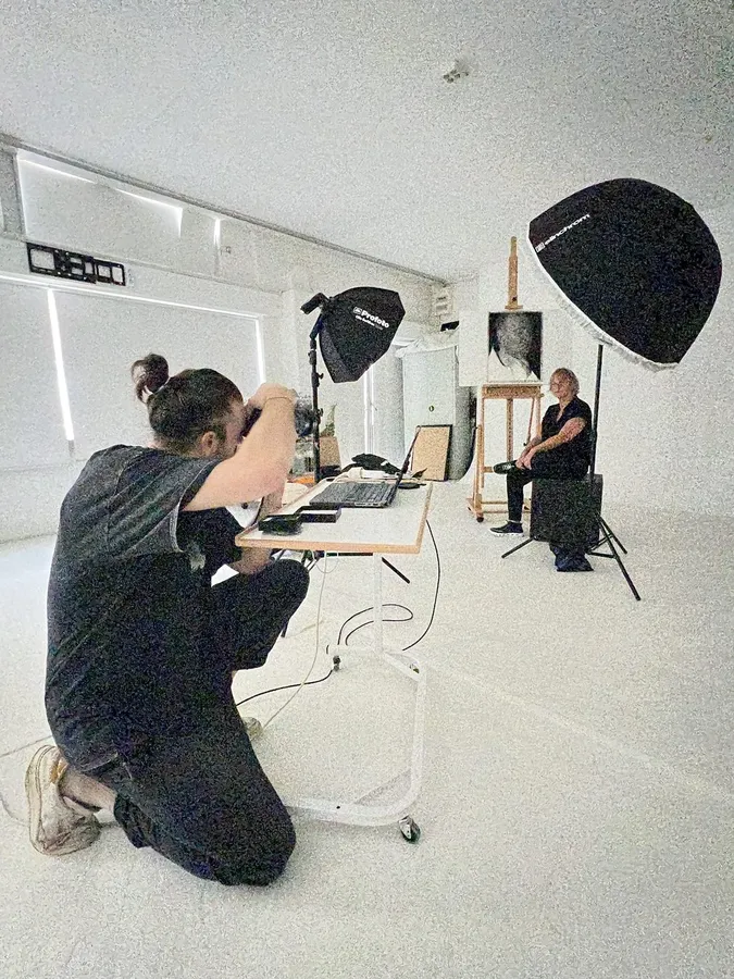 Photographer kneeling at a laptop in a white studio, model and softboxes behind, flash by Kilian Amrehn