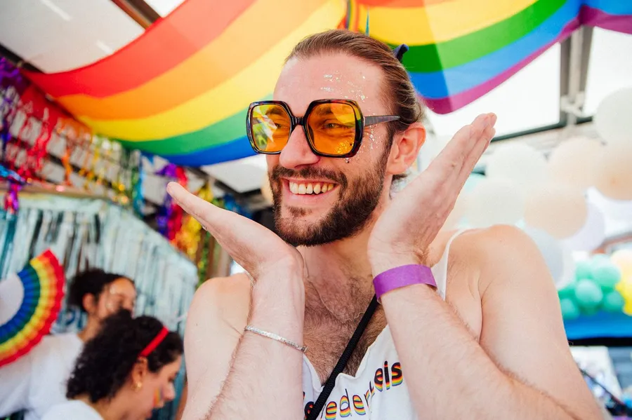 Man with orange sunglasses and glitter laughing under rainbow flags, flash by Kilian Amrehn