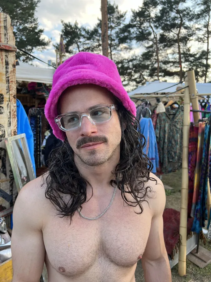 Shirtless man with pink fluffy hat, glasses and curls in front of festival stalls, flash by Kilian Amrehn