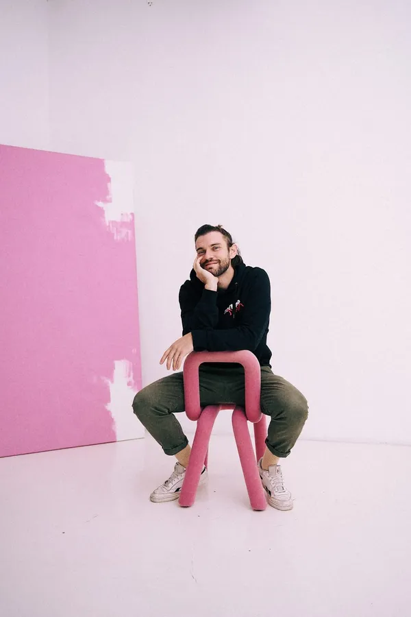 Man sitting backwards on a pink chair in a pink studio, chin on hand, flash by Kilian Amrehn
