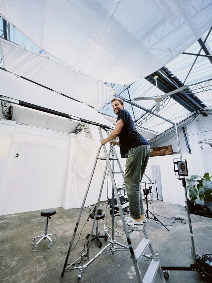 Man laughing on a ladder in a bright hall with glass roof and fabric panels, flash by Kilian Amrehn