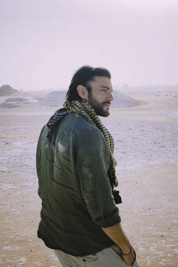Man with a keffiyeh and green jacket looking aside, sandy expanse in haze, flash by Kilian Amrehn