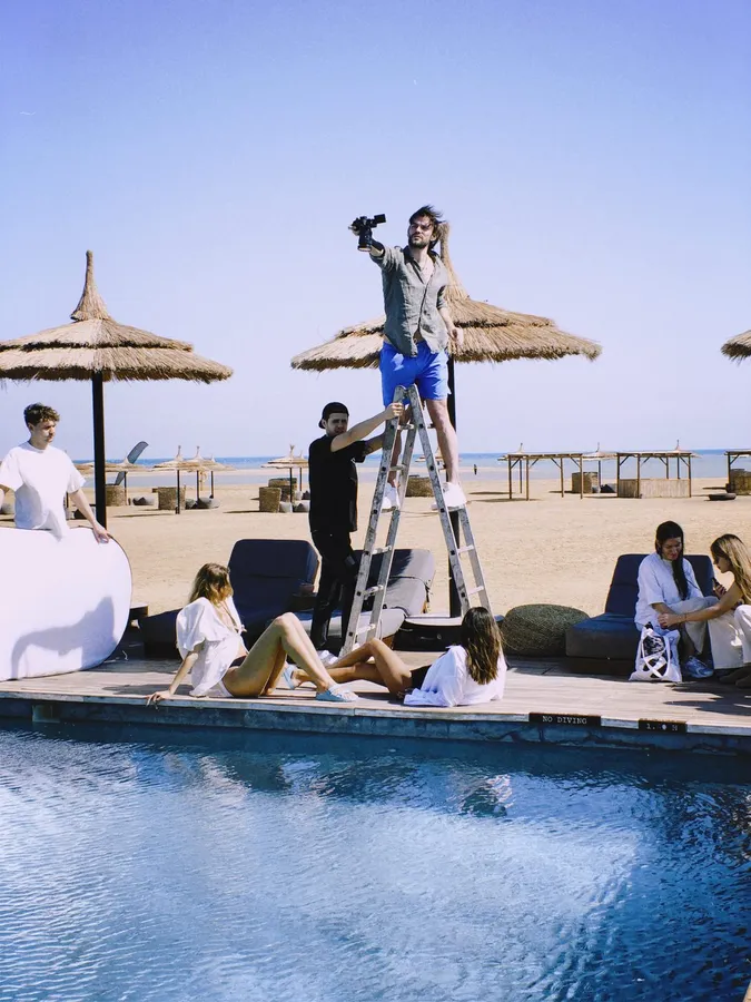 Shoot at a beach pool: photographer on a ladder, models under straw parasols, flash by Kilian Amrehn