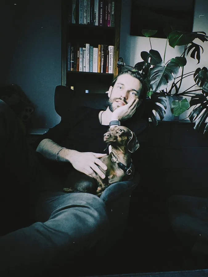 Man slouching in an armchair with a dachshund on his lap, bookshelf and monstera, flash by Kilian Amrehn