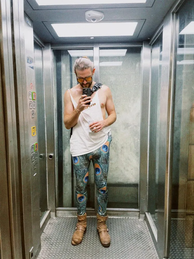 Mirror selfie in a lift: man in colourful leggings and a white tank top, flash by Kilian Amrehn