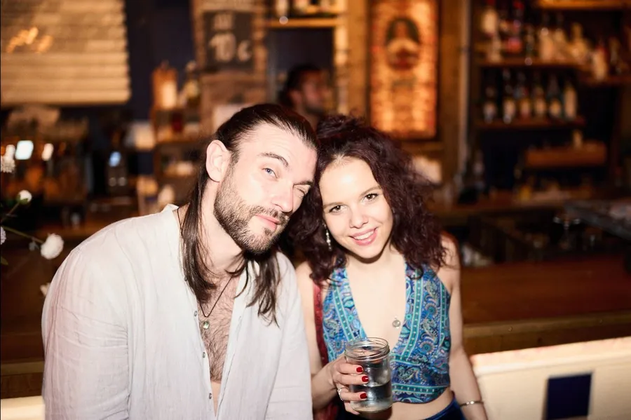 Man with long hair and a smiling woman with a glass in a warm bar, flash by Kilian Amrehn