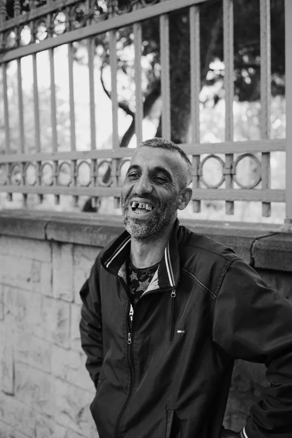 Black and white: laughing man in a jacket in front of an iron fence, Istanbul, flash by Kilian Amrehn