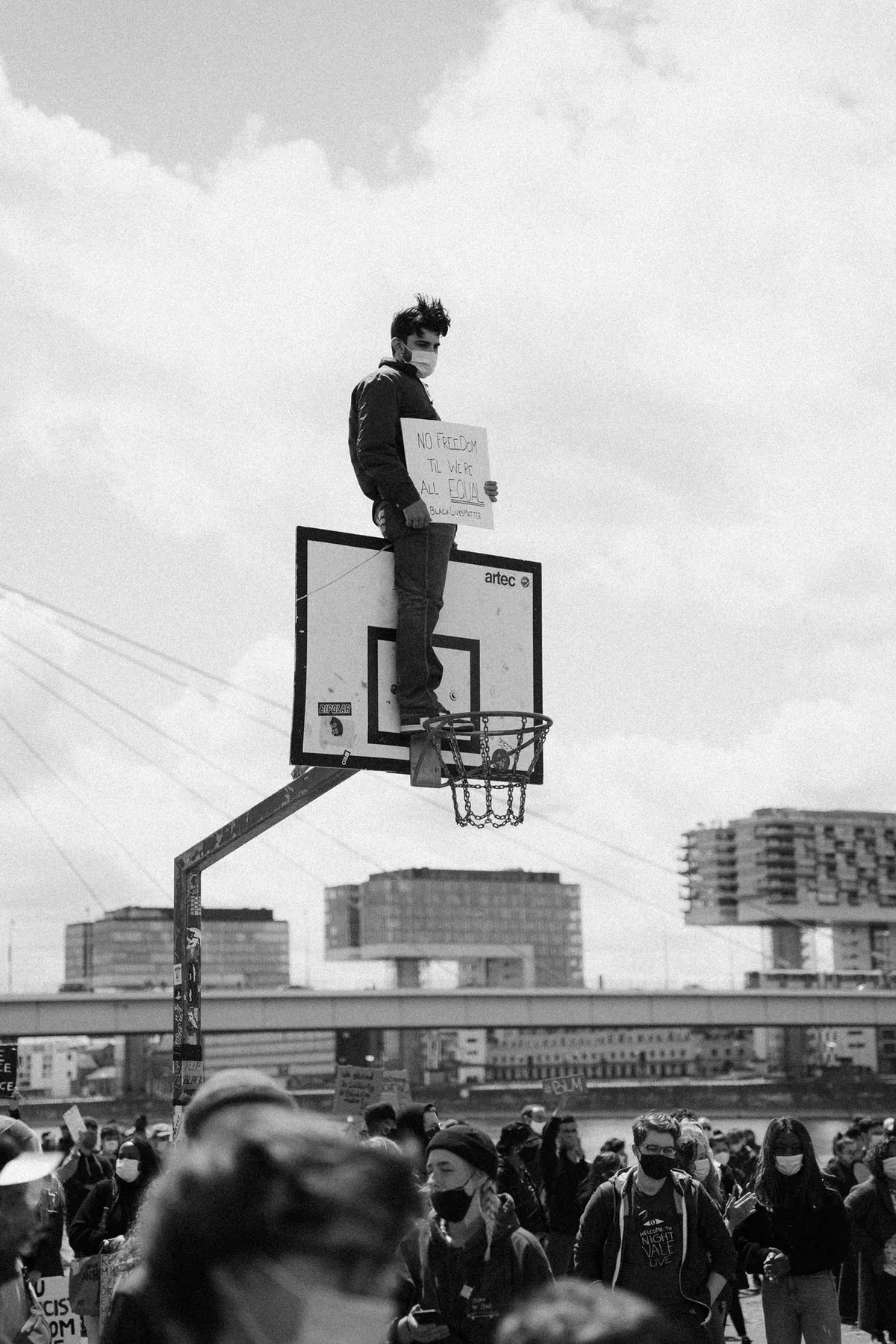 Demonstrant mit Schild steht auf einem Basketballkorb über der Menge, Kranhäuser im Hintergrund, schwarzweiß, Black Lives Matter Köln 2020, Reportage von Kilian Amrehn