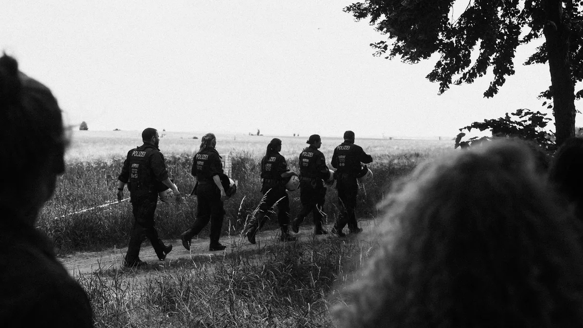 Polizeikette läuft über einen Feldweg, im Vordergrund Köpfe von Zuschauenden, schwarzweiß, Fridays for Future Garzweiler 2019, Reportage von Kilian Amrehn