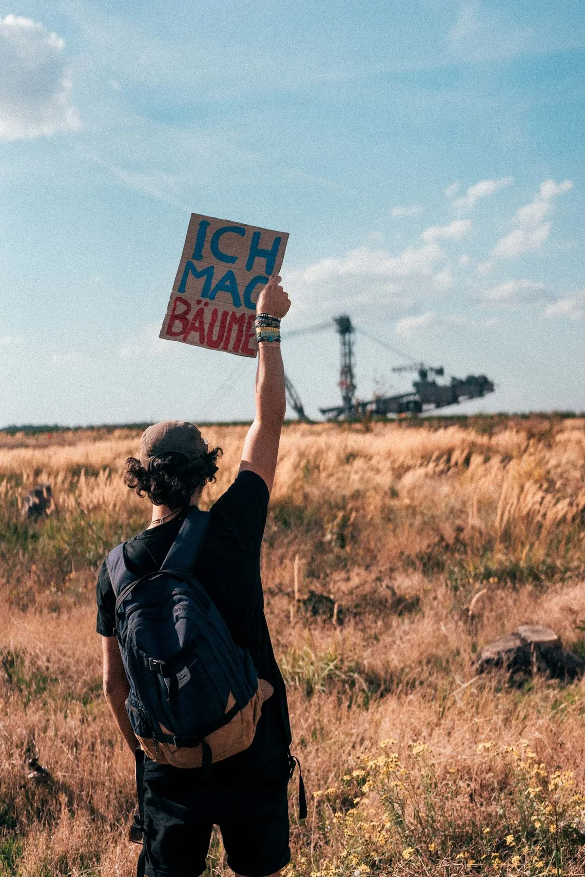 Person mit Rucksack hält Schild Ich mag Bäume vor Feld und Schaufelradbagger, blauer Himmel, Hambacher Forst 2018, Reportage von Kilian Amrehn