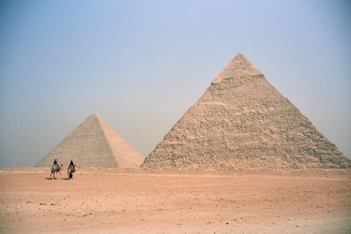 Zwei Pyramiden mit Kamelreitern davor im Wüstensand, dunstiger blauer Himmel, Kairo 2023, Reportage von Kilian Amrehn