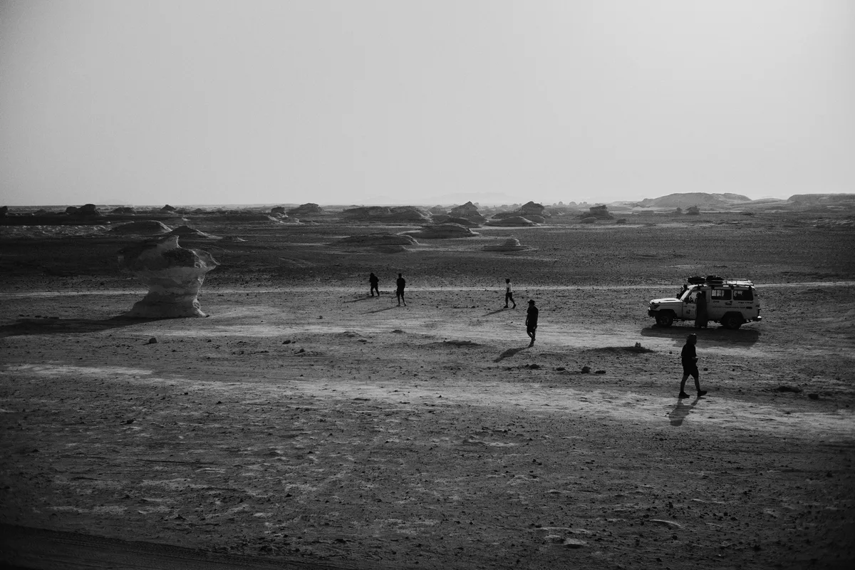 Kleine Menschen und Geländewagen zwischen Felsformationen in weiter Wüste, schwarzweiß, Ägypten 2023, Reportage von Kilian Amrehn