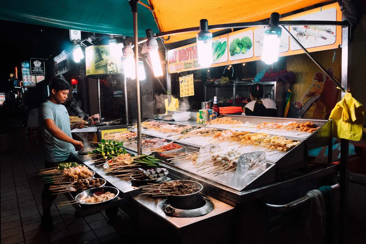 Nachtmarktstand mit Spießen unter grellen Lampen, Mann grillt, orangefarbenes Vordach, Malaysia 2017, Reportage von Kilian Amrehn