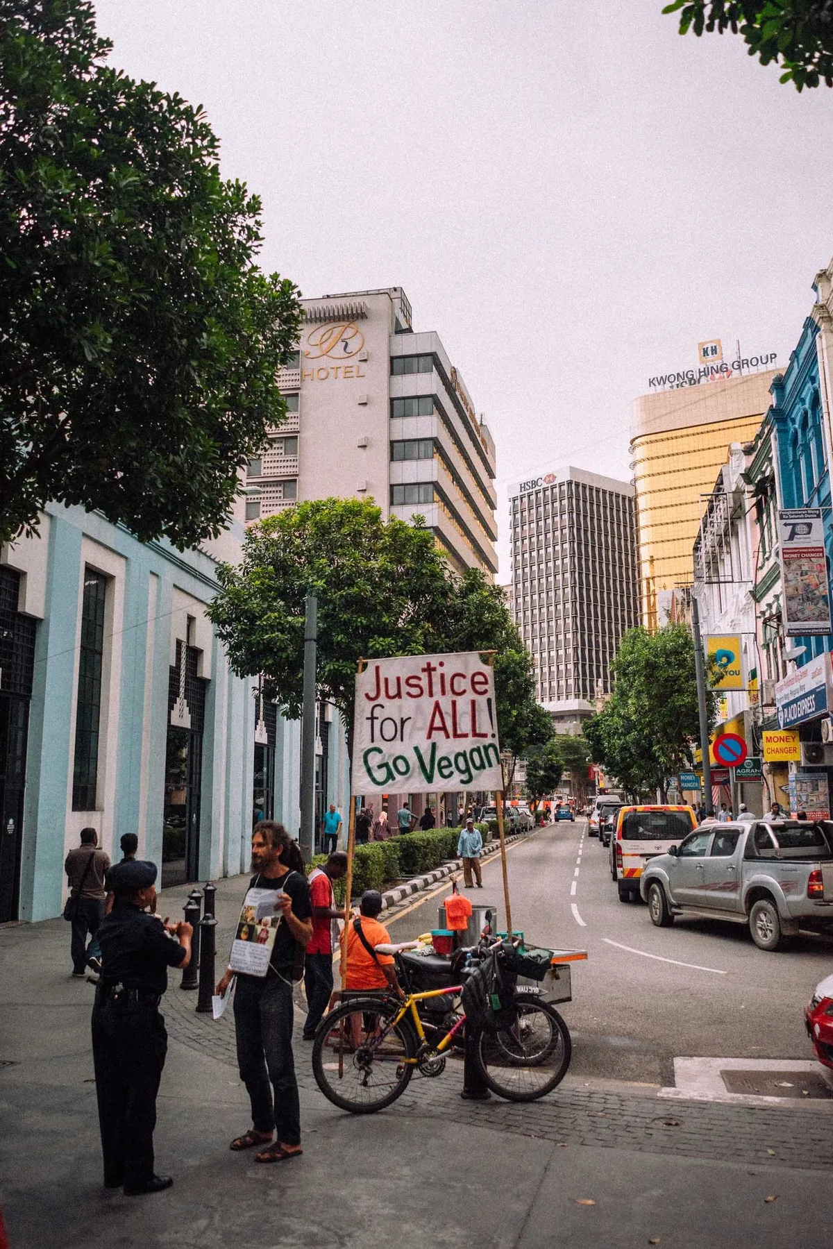 Straßenecke mit Mann und Schild Justice for All Go Vegan neben Polizistin und Fahrrad, Hochhäuser, Malaysia 2017, Reportage von Kilian Amrehn