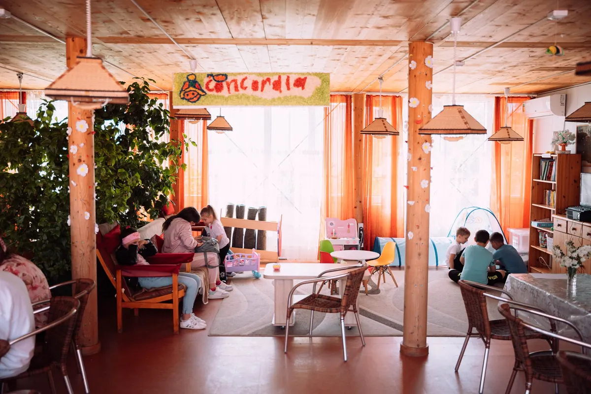 Heller Gemeinschaftsraum mit Holzsäulen und orangen Vorhängen, Kinder spielen auf dem Teppich, Flüchtlingsunterkunft in Moldau 2022, Reportage von Kilian Amrehn